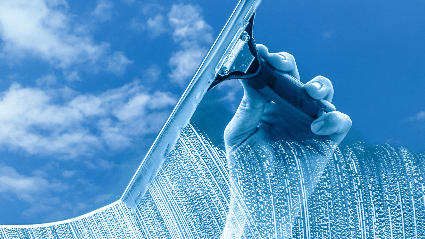 Mão limpando cobertura de vidro com rodo e solução de limpeza, céu azul refletido no vidro.